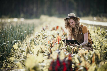 Frau mit Hut, die lächelnd ein digitales Tablet auf einem Bauernhof benutzt - MJRF00313
