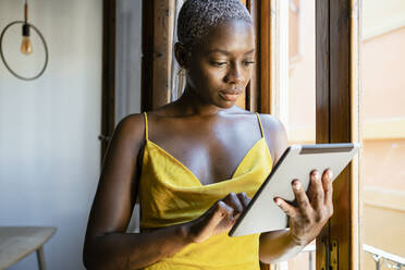 Modischer weiblicher Hipster, der ein digitales Tablet benutzt, während er sich zu Hause ans Fenster lehnt - RCPF00447
