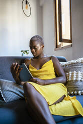 Frau im gelben Kleid benutzt digitales Tablet, während sie zu Hause auf dem Sofa sitzt - RCPF00455