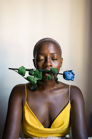 Frau schließt die Augen, während sie mit einer blauen Rose im Mund zu Hause an der Wand sitzt, lizenzfreies Stockfoto