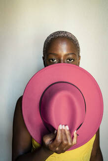 Confident woman holding pink hat in front of face against white wall at home - RCPF00481