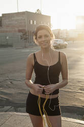 Smiling woman holding jump rope standing on sidewalk in city at sunset - AJOF00725