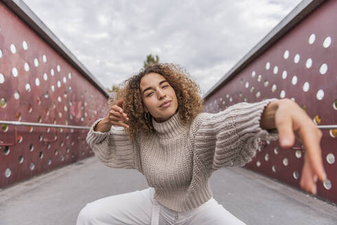 Lächelnde junge Frau tanzt, während sie auf einer Brücke gegen den Himmel hockt - MEF00019