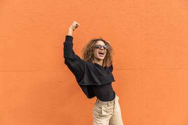Fröhliche Frau mit erhobenen Armen an einer orangefarbenen Wand stehend - MEF00036