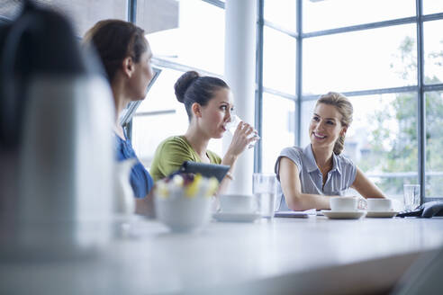 Smiling businesswoman sitting by female colleague drinking water at conference table in office - AJOF00730
