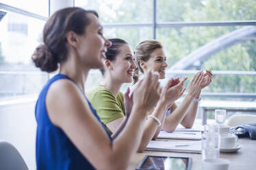 Happy female colleagues applauding while sitting in board room during meeting in office - AJOF00732