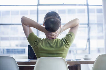 Müde Geschäftsfrau sitzt mit den Händen im Nacken am Schreibtisch im Büro - AJOF00736