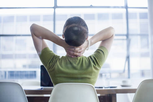 Tired businesswoman sitting with hands behind neck at desk in office - AJOF00736