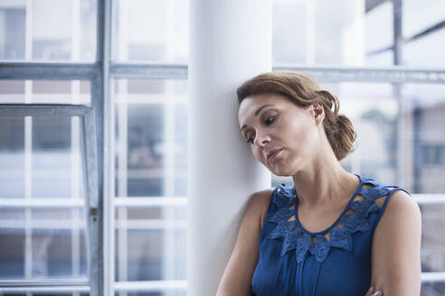 Pensive businesswoman leaning on column in office - AJOF00738