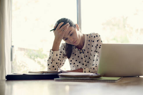 Müde Geschäftsfrau sitzt mit dem Kopf in den Händen bei der Arbeit im Büro - AJOF00740