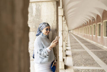 Young woman wearing headscarf using mobile phone while leaning on pillar - JCCMF00034