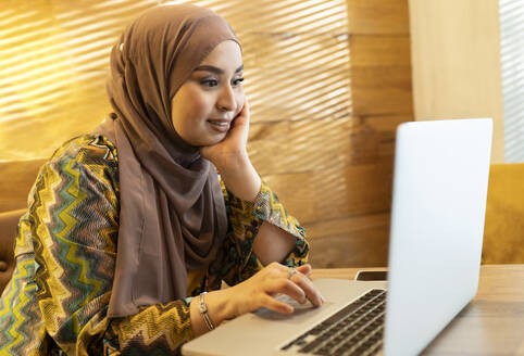 Junge Frau mit Hidschab, die mit der Hand am Kinn sitzt, während sie in einem Café am Laptop arbeitet - JCCMF00039