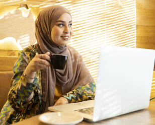 Frau trägt Hijab und trinkt Kaffee, während sie in einem Café sitzt - JCCMF00040