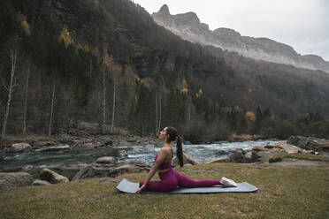 Active woman practicing yoga on mat by river and mountains at Ordesa National Park, Huesca, Spain - ACPF00940