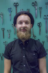 Smiling young male barber standing against scissors hanging on turquoise wall  - AJOF00747