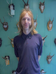 Blond male barber standing against animal skulls and antlers on turquoise wall - AJOF00748