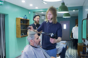 Männlicher Friseur, der einen Haartrockner auf das Haar eines Kunden anwendet - AJOF00754