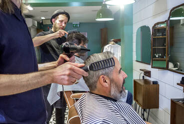 Männlicher Friseur, der einen Fön benutzt, während er die Haare eines Kunden kämmt, in einem Friseursalon - AJOF00759