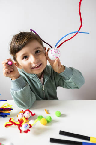 Smiling girl showing creative toys made of pipe cleaners and styrofoam ball against wall at home stock photo