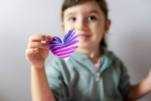 Glückliches Mädchen hält handgefertigtes Herz mit Origami gegen die Wand - GEMF04430