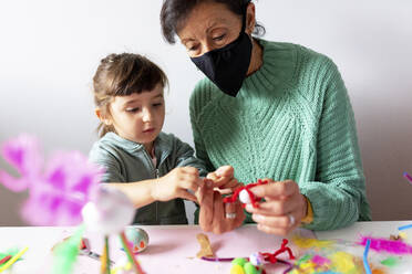 Niedliche Enkelin mit Großmutter beim Basteln von kreativem Spielzeug aus Pfeifenreinigern und Pompons während COVID-19 - GEMF04433