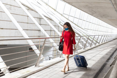 Ganzkörper-Touristin mit Schutzmaske, die mit ihrem Gepäck in der Halle des Flughafens spazieren geht und Nachrichten auf ihrem Smartphone liest, während sie auf ihren Flug wartet, während der Coronavirus-Pandemie - ADSF18428