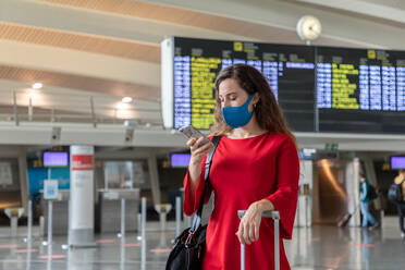 Serious female traveler in mask standing with suitcase on background of departure board and browsing cellphone while waiting for flight - ADSF18429