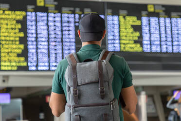 Rückenansicht eines nicht erkennbaren männlichen Reisenden mit Rucksack, der den Zeitplan auf der Abflugtafel überprüft, während er im internationalen Flughafen steht und auf seinen Flug wartet - ADSF18430