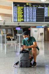 Ruhiger männlicher Tourist hockt an der Abflugtafel im Flughafen und chattet mit seinem Smartphone in den sozialen Medien, während er auf seinen Flug wartet - ADSF18431