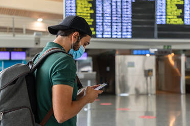 Rückenansicht eines ruhigen männlichen Touristen, der an der Abflugtafel im Flughafen steht und über sein Smartphone in den sozialen Medien chattet, während er auf seinen Flug wartet - ADSF18432
