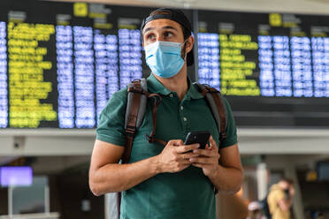 Calm male tourist standing against departure board in airport and chatting on social media via smartphone while waiting for flight - ADSF18433