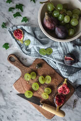 Draufsicht auf köstliche Trauben und Feigen, die auf einem Marmortisch mit Schneidebrett und Küchentuch angeordnet sind - ADSF18434