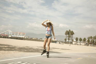 Young woman holding hat while inline skating by beach on sunny day - AJOF00763