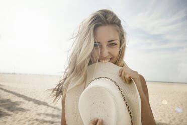 Verspielte junge Frau mit blondem Haar beißt Hut am Strand an einem sonnigen Tag - AJOF00770
