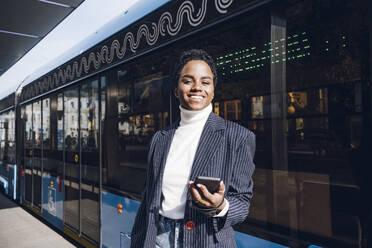 Lächelnde Geschäftsfrau, die ihr Mobiltelefon an einem sonnigen Tag gegen einen Bus hält - VYF00289