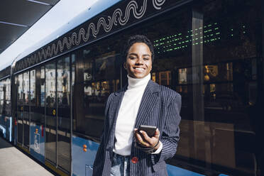 Smiling businesswoman holding mobile phone against bus on sunny day - VYF00289