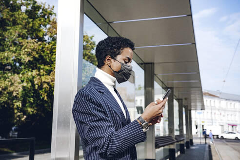 Young businesswoman in protective face mask using mobile phone at bus stop - VYF00290