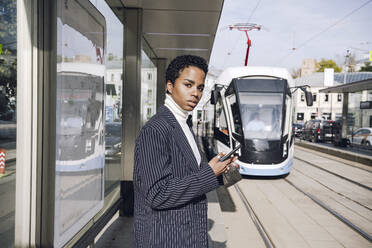 Young businesswoman with mobile phone waiting for bus at stop on sunny day - VYF00294