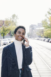 Happy businesswoman looking away while talking on smart phone in city during sunny day - VYF00299