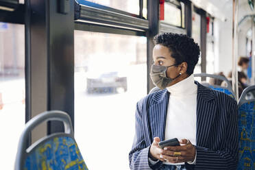 Junge Geschäftsfrau mit Mobiltelefon, die durch das Fenster schaut, während sie in einem Bus während COVID-19 pendelt - VYF00301