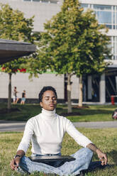 Junge Geschäftsfrau mit Laptop beim Meditieren im Park an einem sonnigen Tag - VYF00317