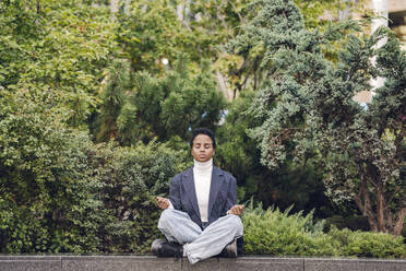 Junge Geschäftsfrau macht Yoga, während sie auf einer Stützmauer gegen Pflanzen sitzt - VYF00338