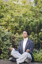 Young businesswoman meditating while sitting on retaining wall against plants - VYF00339