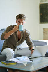 Businessman drinking coffee while working on laptop at home - KIJF03382