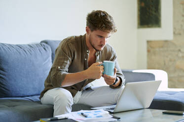 Young male entrepreneur holding coffee mug while working on laptop in living room - KIJF03383