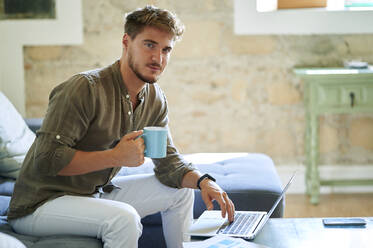 Male freelancer holding coffee mug while sitting by laptop at home - KIJF03384