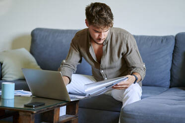 Young male freelancer reading documents while sitting on sofa with laptop at home - KIJF03386