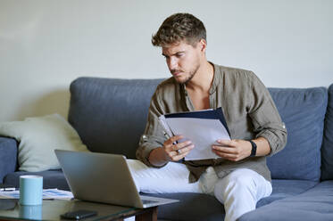 Businessman working on laptop while holding paper in living room - KIJF03387