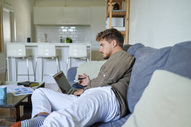 Businessman using laptop while lying on sofa at home - KIJF03394