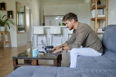 smiling male entrepreneur working at home with laptop while sitting on sofa in living - KIJF03395